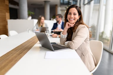 Person sat at a desk with an open laptop. They are turned towards the camera smiling.